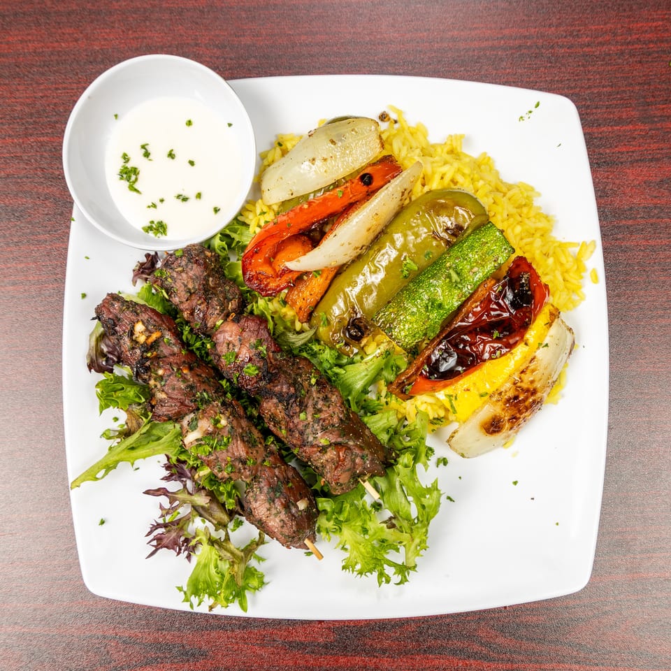 Grilled beef skewers, roasted vegetables, yellow rice, salad greens, and creamy dipping sauce served on a white plate.