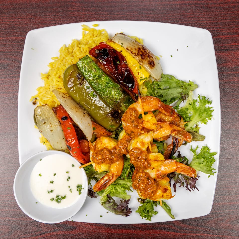 Platter of grilled shrimp skewers, vegetables, yellow rice, salad greens, and creamy dipping sauce on a white plate.