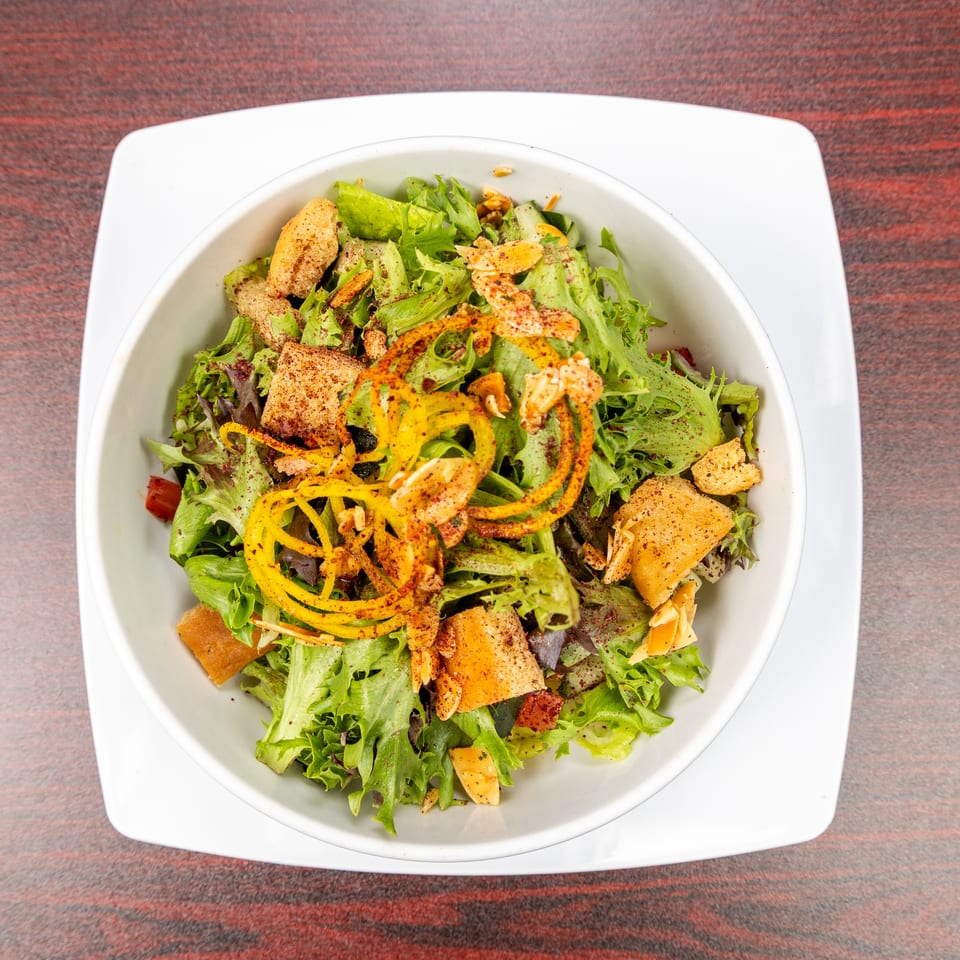 Overhead view of a mixed green salad with orange vegetable strips, croutons, sliced almonds, and spices in a white bowl.