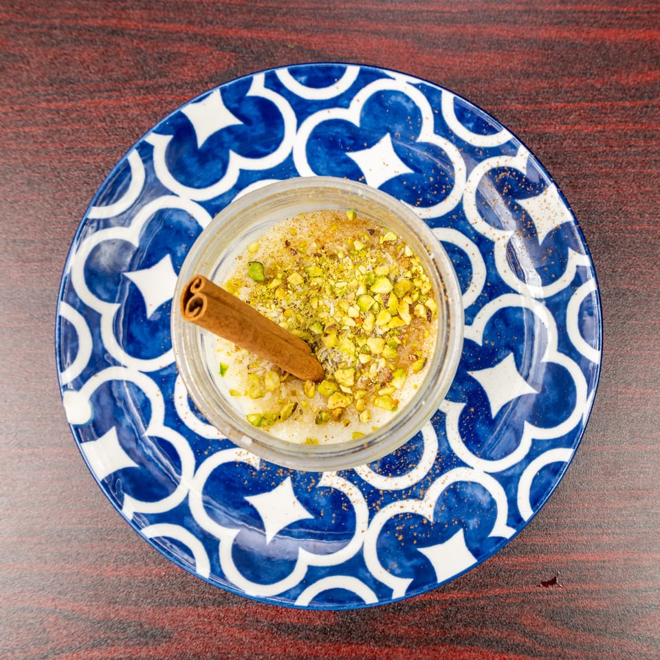 Creamy dessert topped with chopped pistachios and a cinnamon stick, served in a glass bowl on a blue patterned plate.